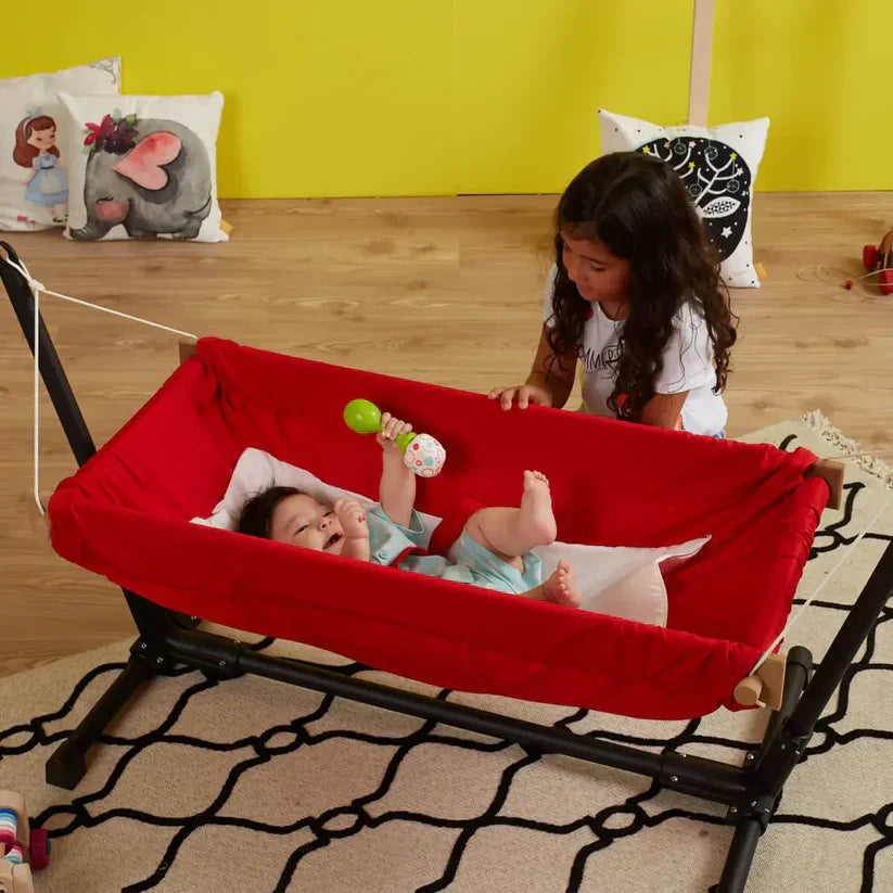 Kırmızı bebek hamağında yatan bebek ve yanında duran çocuk – bebek hamaklarının güvenli kullanımı